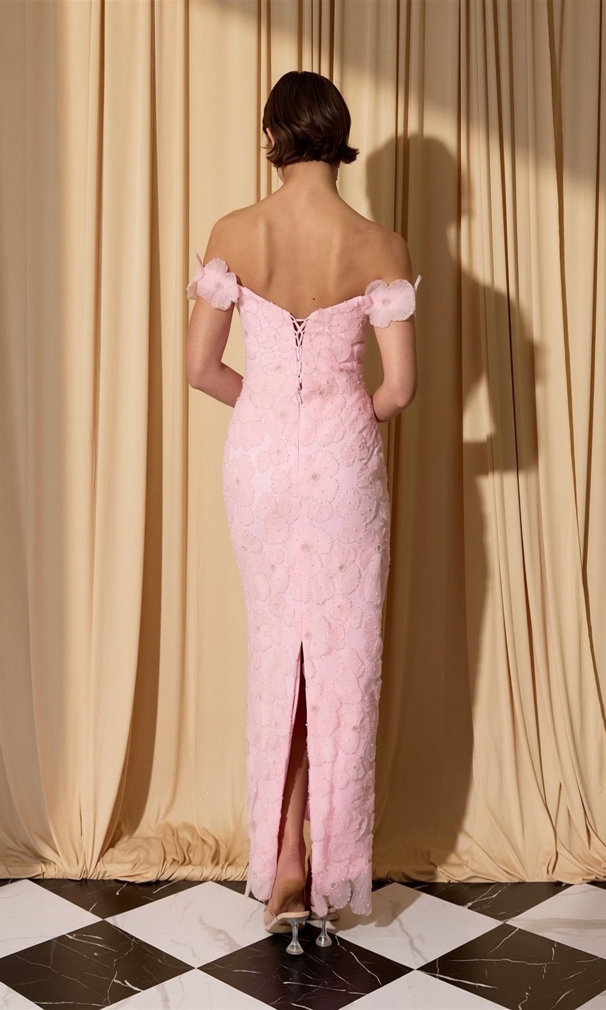 A woman stands on a black and white tiled floor, facing away, wearing the Eureka EK205 long prom dress in pale pink lace with 3D floral appliqué, off the shoulder design, and back slit. Cream curtains are draped behind her.